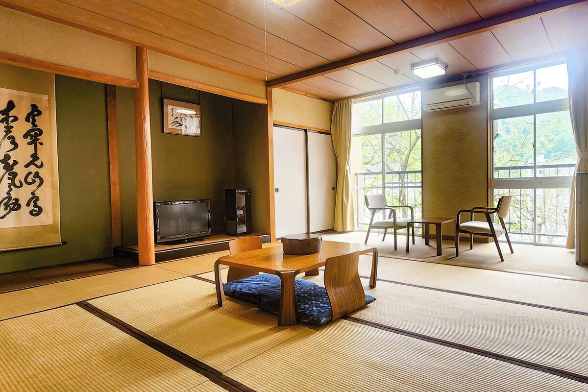 A traditional Japanese room with tatami flooring, a low table, chairs, a television, and a large window with a view of greenery.