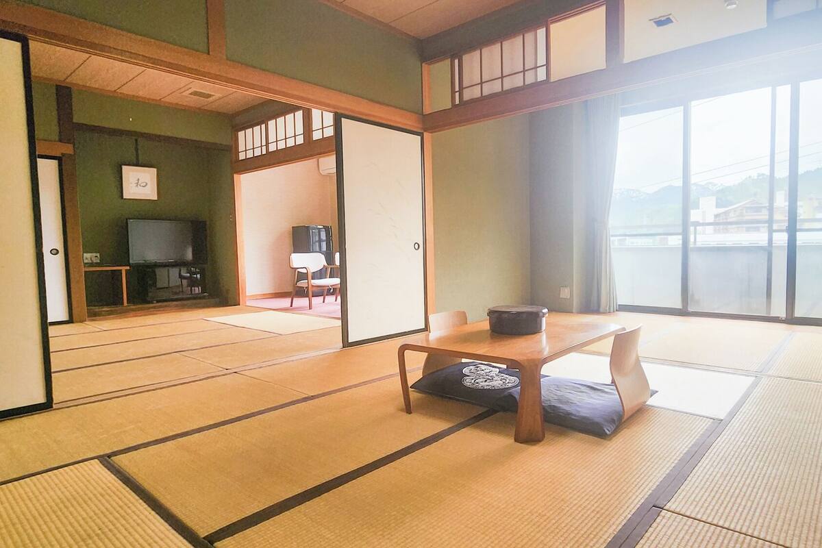 A traditional Japanese room with tatami flooring, a low table with a cushion, a television, and a view of a body of water.