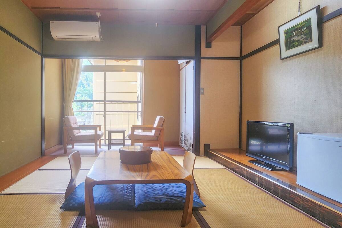 A traditional Japanese room with tatami flooring, a low table with cushions, a television, and a sliding door leading to a balcony.