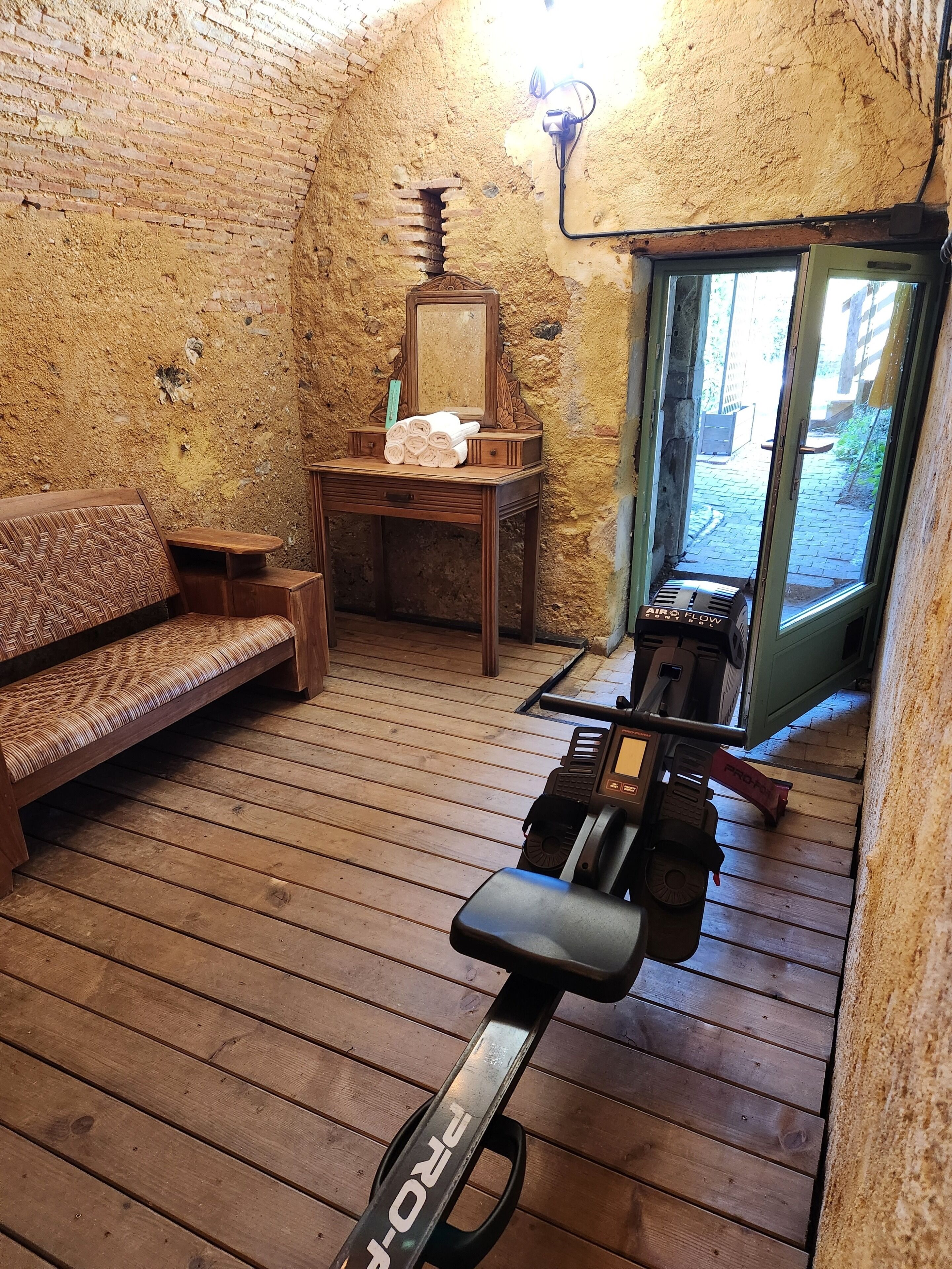Une pièce rustique avec un banc en bois, une petite table avec des serviettes, un miroir et un vélo d’appartement.