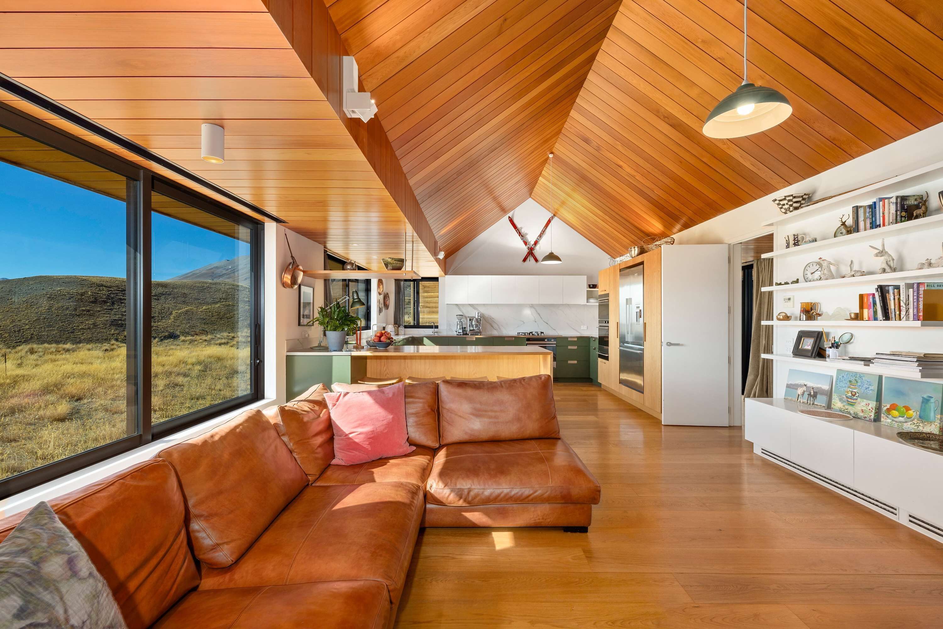 A modern interior with a wooden ceiling, a leather sofa, a kitchen area, and a large window overlooking a landscape.