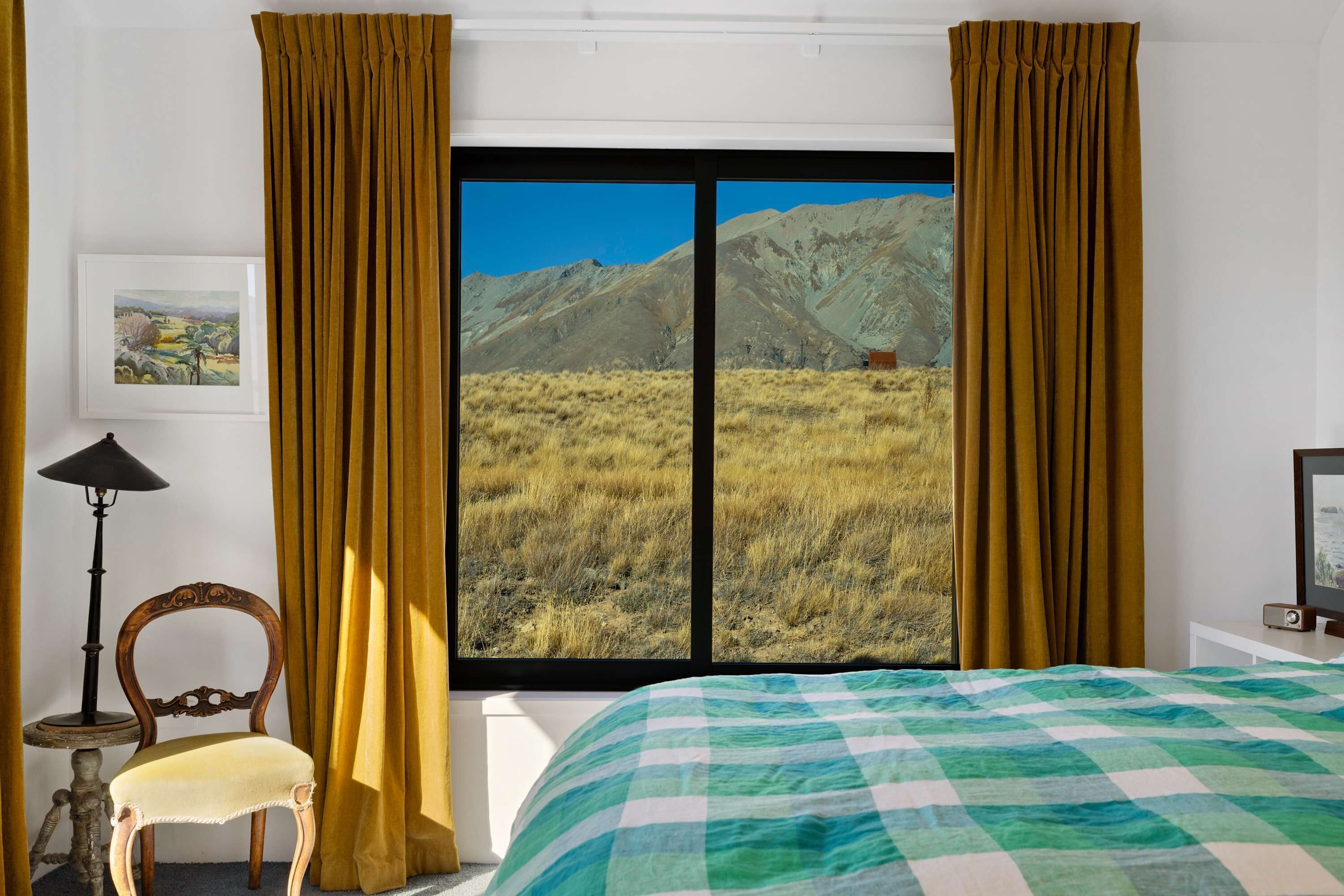 Una habitación con una cama, una silla, una lámpara, un cuadro enmarcado y un ventanal con cortinas.