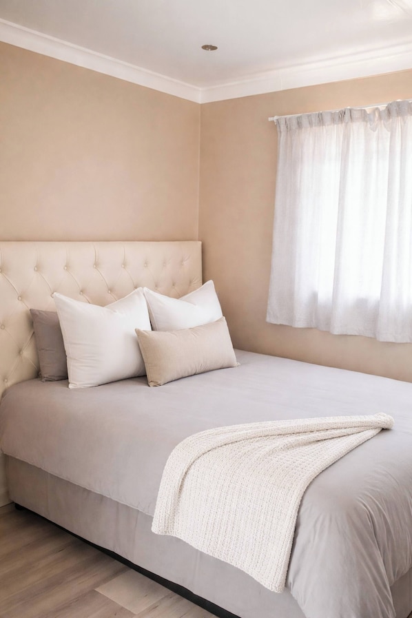 Un dormitorio con cabecera capitonada, cama con ropa blanca, una manta blanca y una ventana con cortinas de gasa.