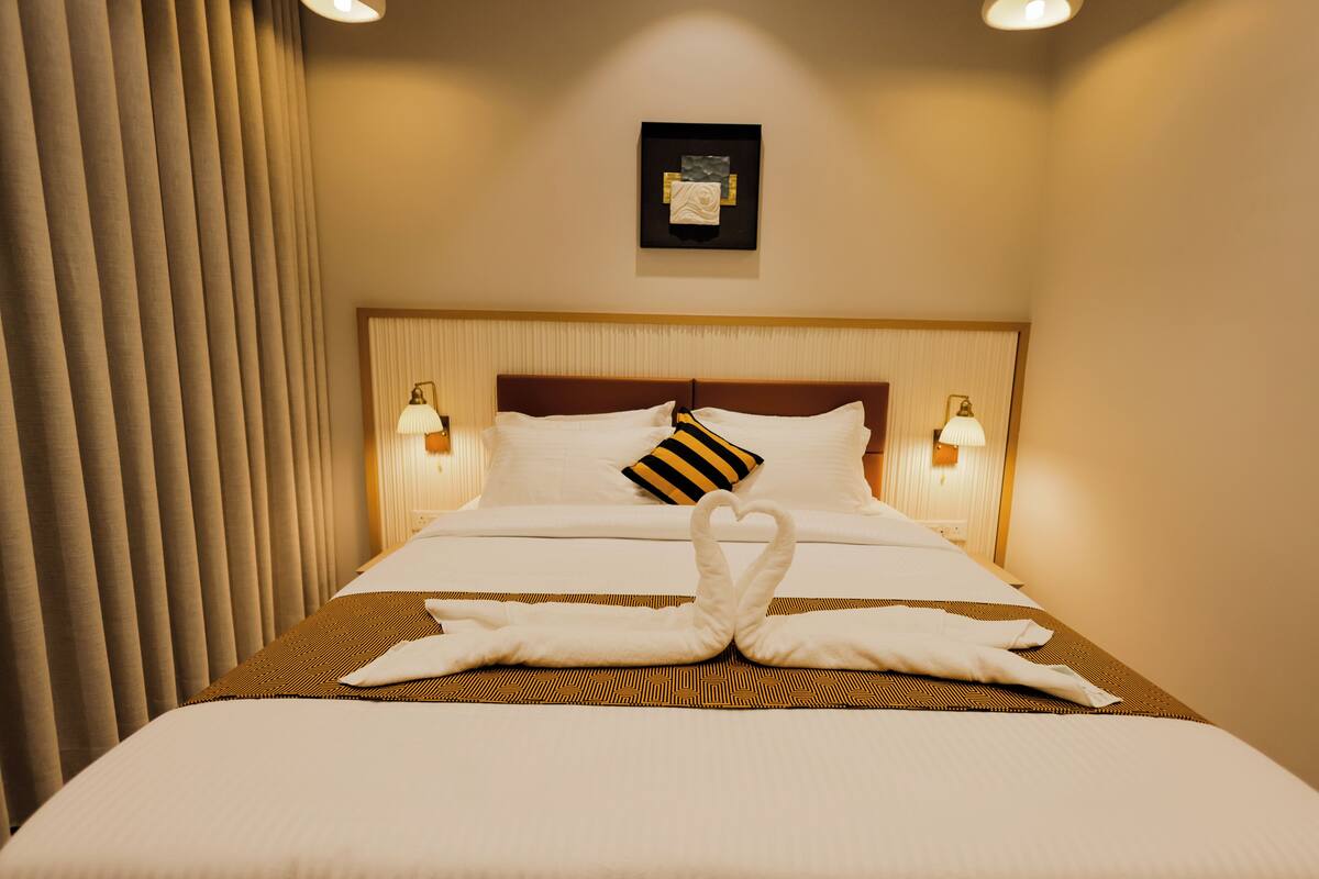 Ein Hotelzimmer mit einem ordentlich bezogenen Bett, einem Kopfteil mit einem Bild, zwei Nachttischlampen und einem Fenster mit Vorhängen.