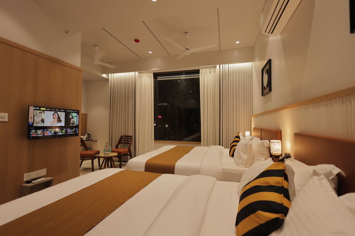 Ein Hotelzimmer mit zwei Betten, einem an der Wand befestigten Fernseher, einem Fenster mit Vorhängen sowie einem Nachttisch mit Lampe.