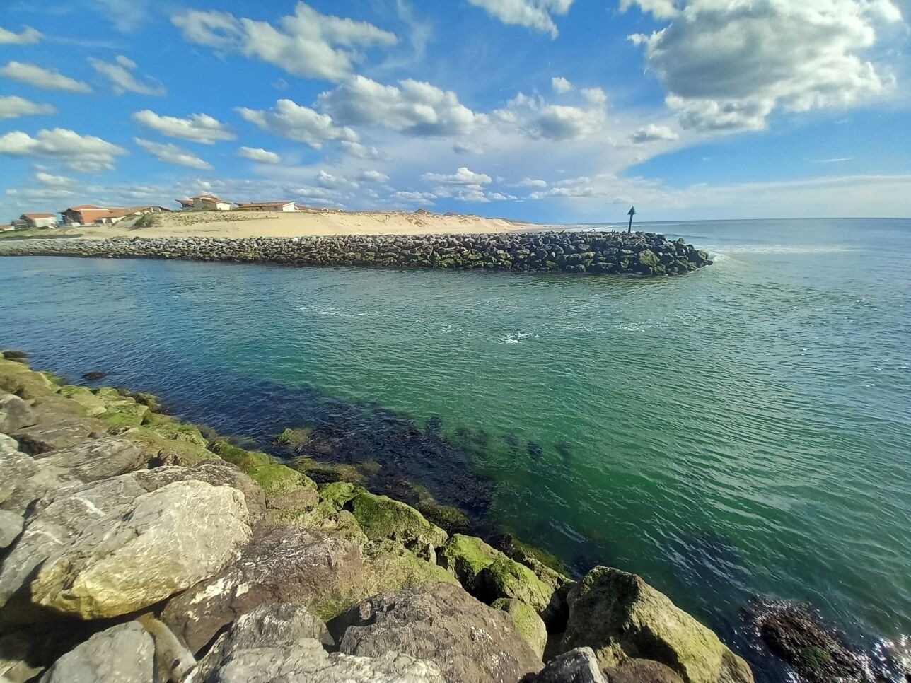 A coastal scene with a rocky shoreline, clear water, and a distant sandy area with buildings.