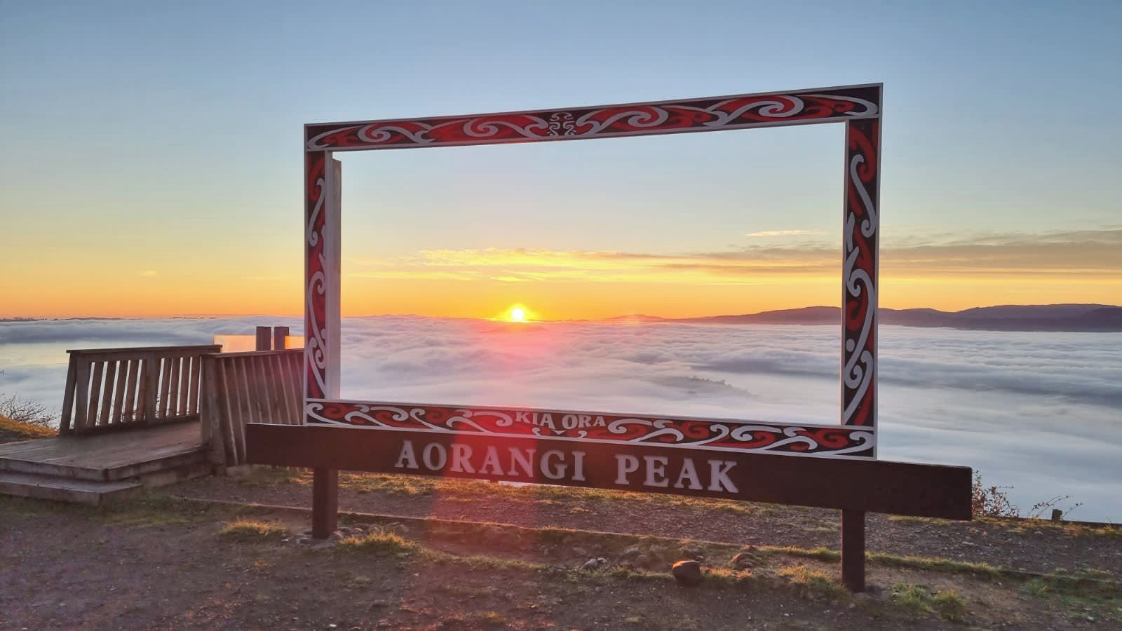 Un cartello di legno con la scritta 'Aorangi Peak' sullo sfondo di un'alba o tramonto.