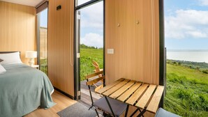 A bedroom with a bed, a wooden table, and a view of a grassy field and water.