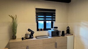 Une petite cuisine équipée d’un four à micro-ondes, d’une machine à café et d’un petit réfrigérateur. Il y a une fenêtre avec des stores, une plante en pot et un meuble avec des tiroirs.