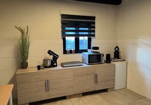 A kitchenette with a microwave, coffee maker, and a small refrigerator. There is a window with blinds, a potted plant, and a cabinet with drawers. - Will's Ranch - Guesthouse (Heibloem)