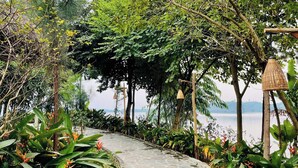 A stone pathway leading through a lush garden with various plants and trees, a pergola structure, and a view of a body of water. - The L'anmien Bui Vien (Ho Chi Minh City)