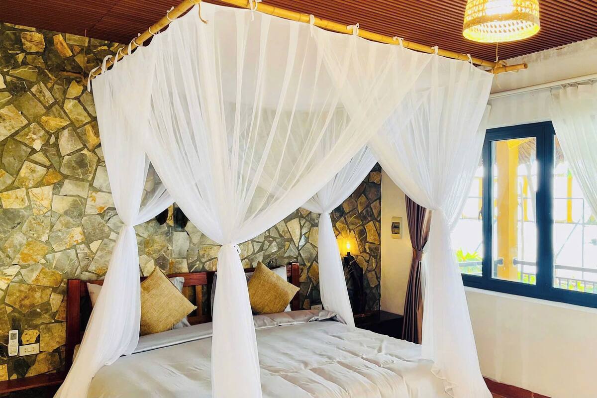 Une pièce avec un mur en pierre, un lit recouvert de draps blancs, un lit à baldaquin et une fenêtre avec des rideaux.