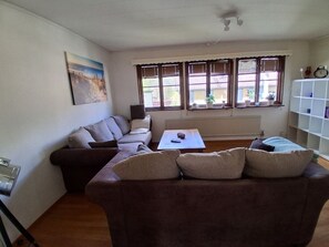 A living room with a sectional sofa, a coffee table, and a radiator. There are windows with blinds and a painting on the wall.