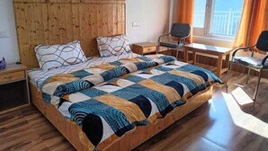 A bedroom with a wooden headboard, a bed with patterned bedding, a desk, two chairs, and a window with curtains. - Hotel Satya (Chamba)