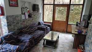 A living room with a sofa, a coffee table, a wooden door, and a small kitchen area with a counter and potted plants. - Hotel Satya (Chamba)