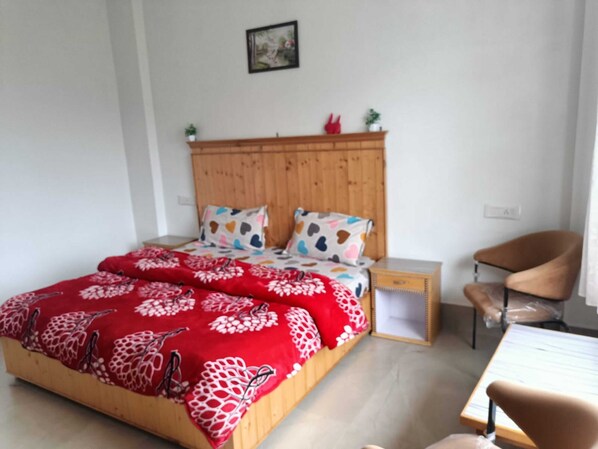 A bedroom with a wooden headboard, a bed with floral and heart-patterned bedding, a bedside table, a chair, and a small table with a chair. - Hotel Satya (Chamba)
