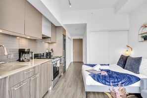 Habitació d'hotel moderna amb cuineta, un llit amb llençol blau i una làmpada de nit.