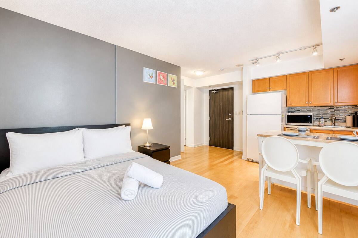 Una habitación de hotel con cama, mesita de noche, lámpara y una kitchenette equipada con microondas, refrigerador y sillas.