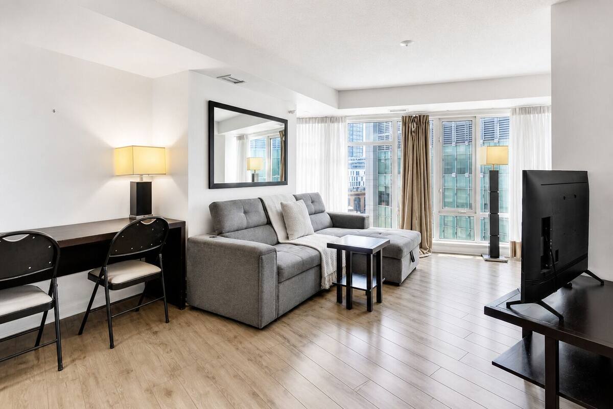 Habitación de hotel moderna con un sofá gris, una mesita pequeña, un televisor, un escritorio con una silla y un ventanal con cortinas.