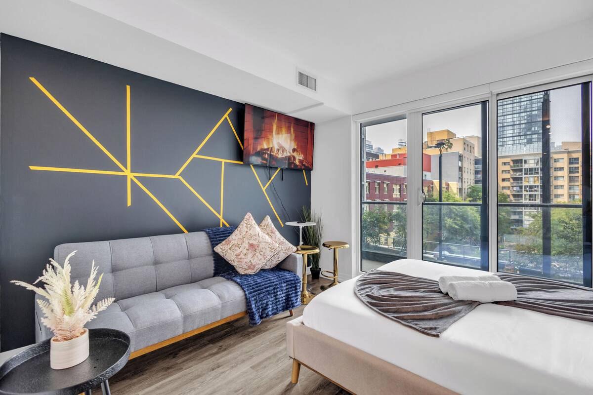 Una habitación de hotel moderna con un sofá gris, una cama con cabecera marrón, un ventanal con vista a la ciudad y un diseño geométrico en la pared.