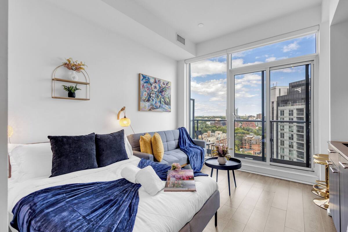 Un dormitorio moderno con una cama grande, un sofá, una mesita y vistas a la ciudad a través de amplios ventanales.