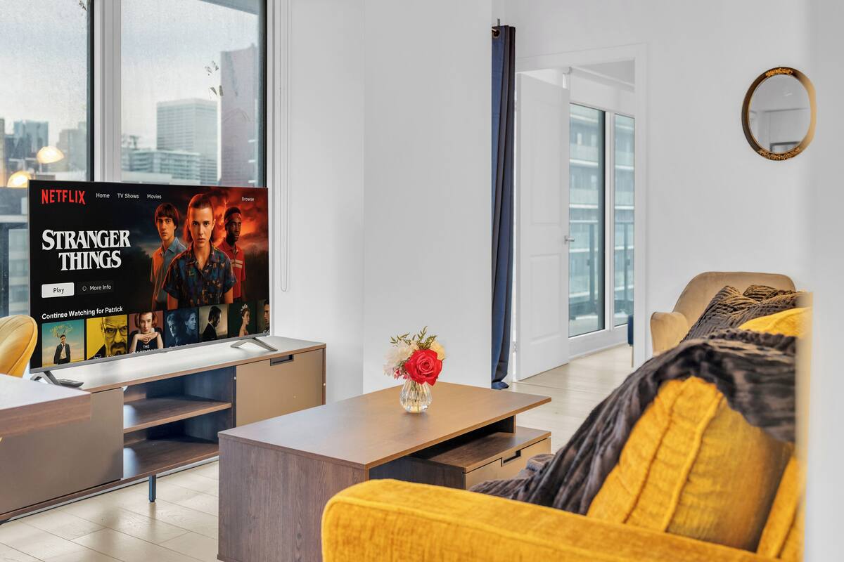 Una sala moderna con un televisor grande mostrando una serie de Netflix, una mesa de centro con un jarrón y un sofá con un cojín amarillo.