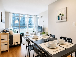 A modern living space with a dining area, a sofa, a TV, and a view of the cityscape through large windows.