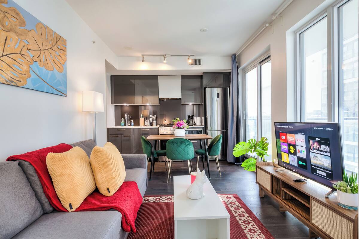 Una sala de estar moderna con un sofá gris, una manta roja y una mesa de centro blanca. Un televisor está sobre un mueble de madera, y hay una zona de comedor con sillas verdes y una mesa de madera.