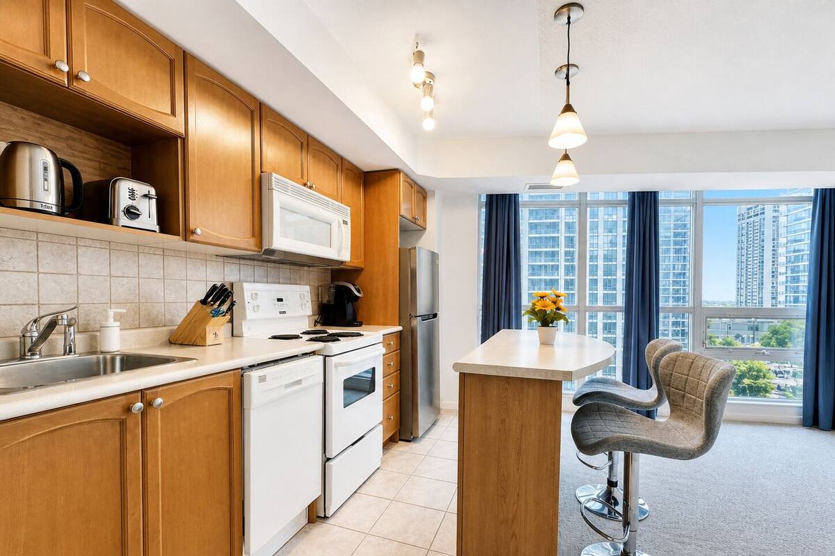 Cocina moderna con armarios de madera, microondas, estufa y un lavabo. Hay una barra con una silla y vistas a la ciudad a través de amplias ventanas.