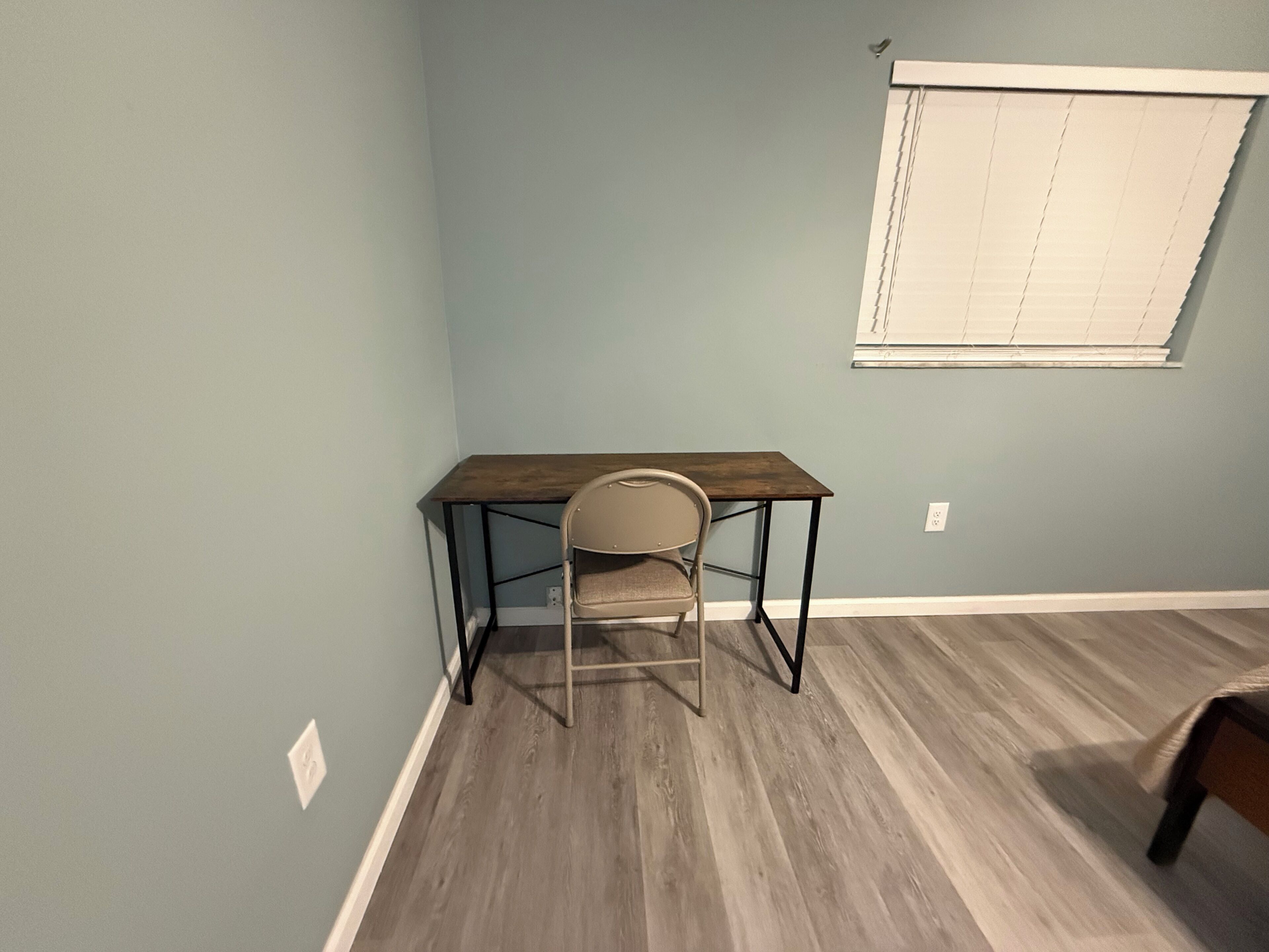 Een kamer met een houten bureau, een stoel, een raam met zonwering en een lichtschakelaar aan de muur.