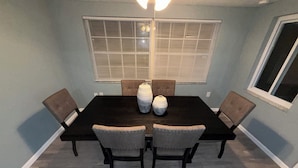 Un coin repas avec une table en bois foncé, six chaises rembourrées et une grande fenêtre équipée de stores.