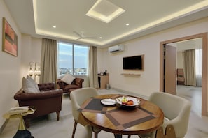 A modern hotel room with a dining area, a sofa set, a flat-screen TV, and a view of the cityscape.
