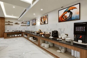 Uma área de pequeno-almoço de hotel moderna, com soalho em mármore, balcão de buffet e máquina de café Nescafé.