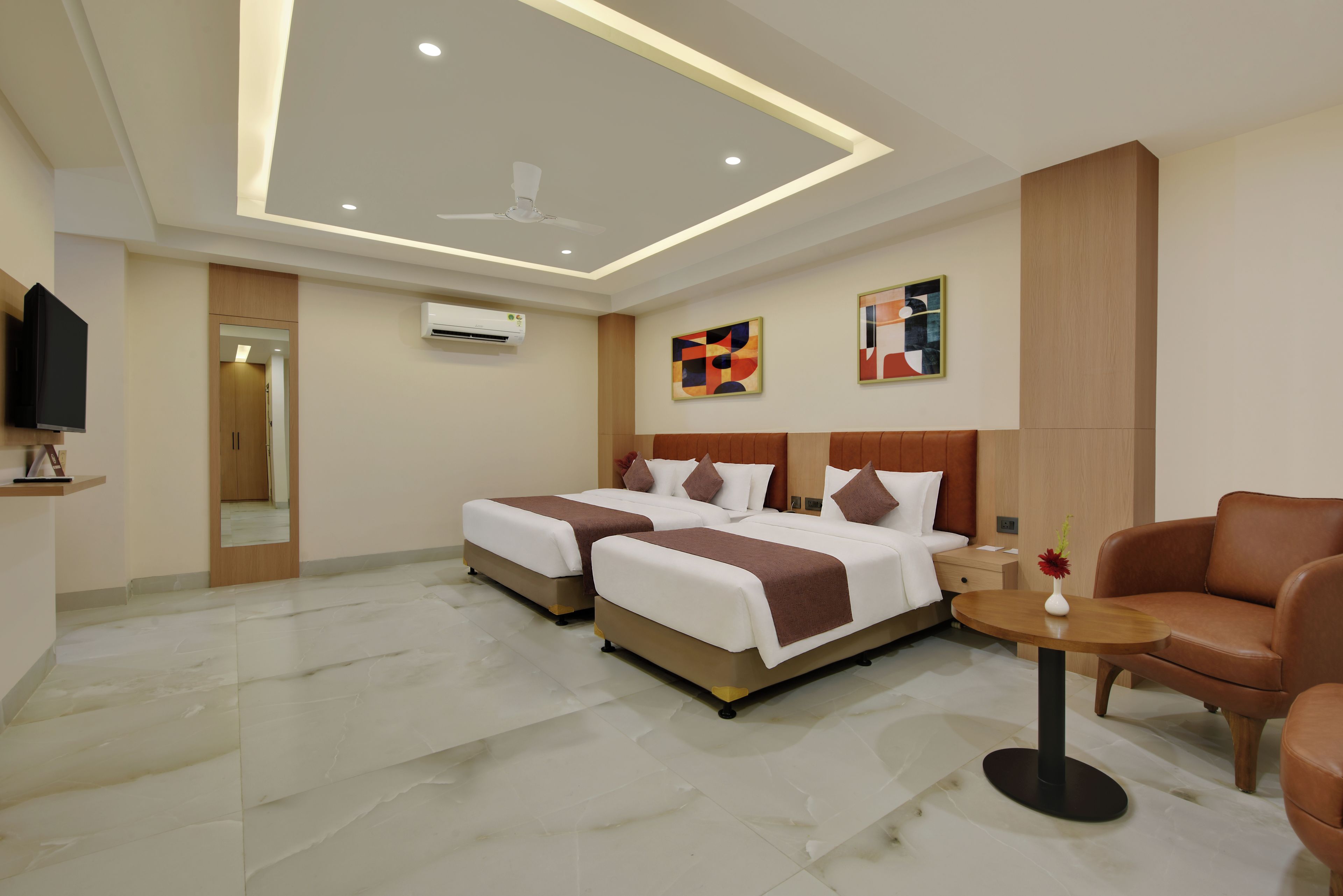 A hotel room with two beds, a TV, an air conditioning unit, and a small table with a vase.