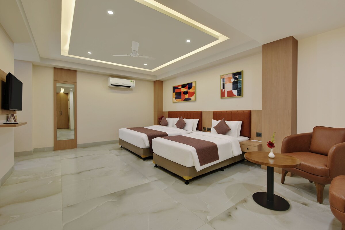 A hotel room with two beds, a TV, an air conditioning unit, and a small table with a vase.
