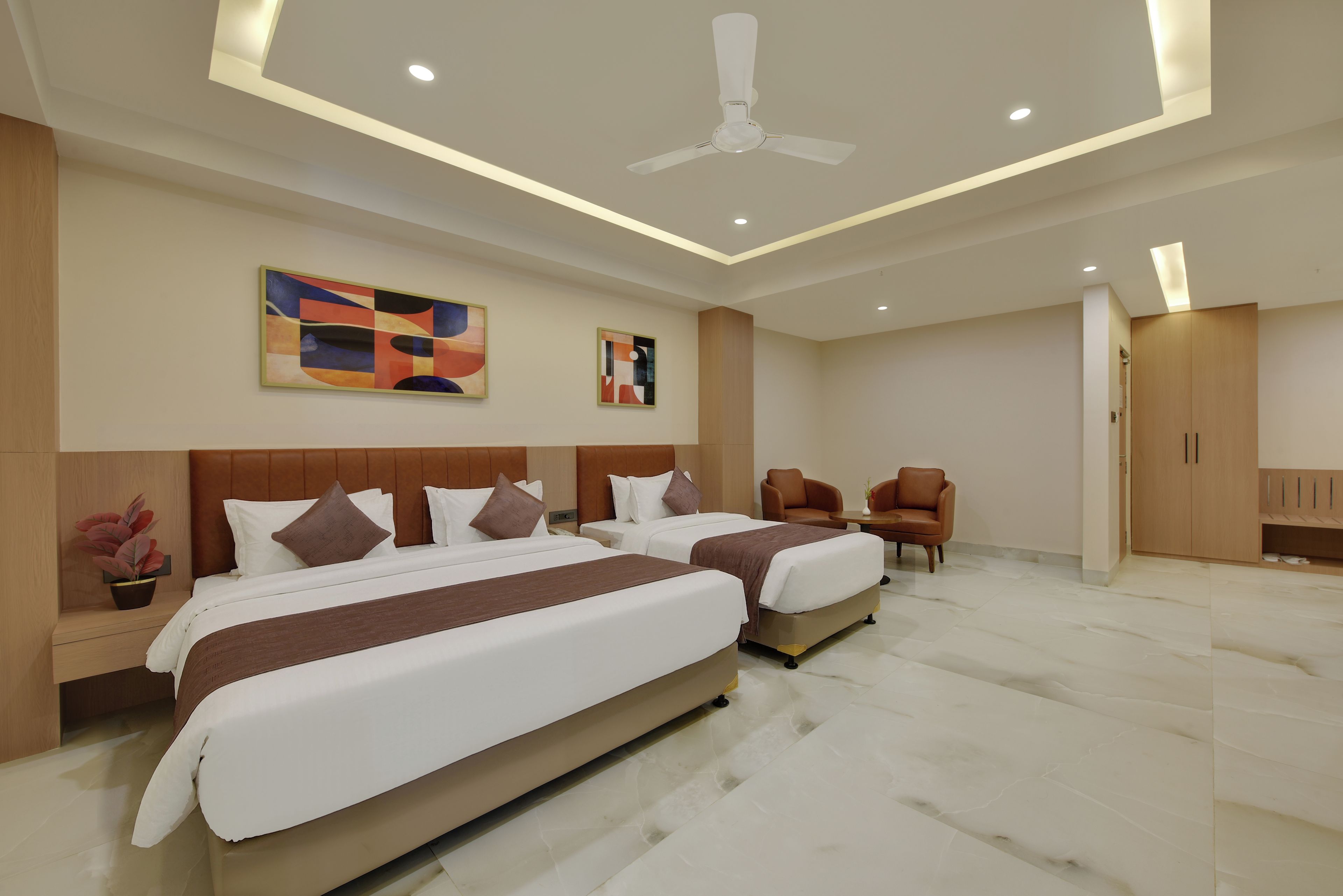 Habitación de hotel moderna con dos camas, ventilador de techo, obras de arte en las paredes y un área de descanso con sillas.