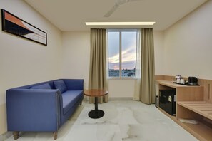 Ein Hotelzimmer mit einem blauen Sofa, einem kleinen runden Tisch, einer Küchenzeile und einem Fenster mit Blick auf die Stadt.