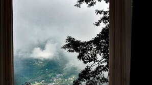 A view through a window with a balcony, featuring a mountainous landscape with greenery and a small settlement below. - Hotel Bella Casa (Gangtok)