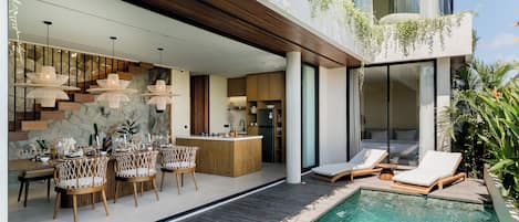A modern outdoor pool area with lounge chairs, a dining area with wicker chairs, and a kitchen visible through glass doors.