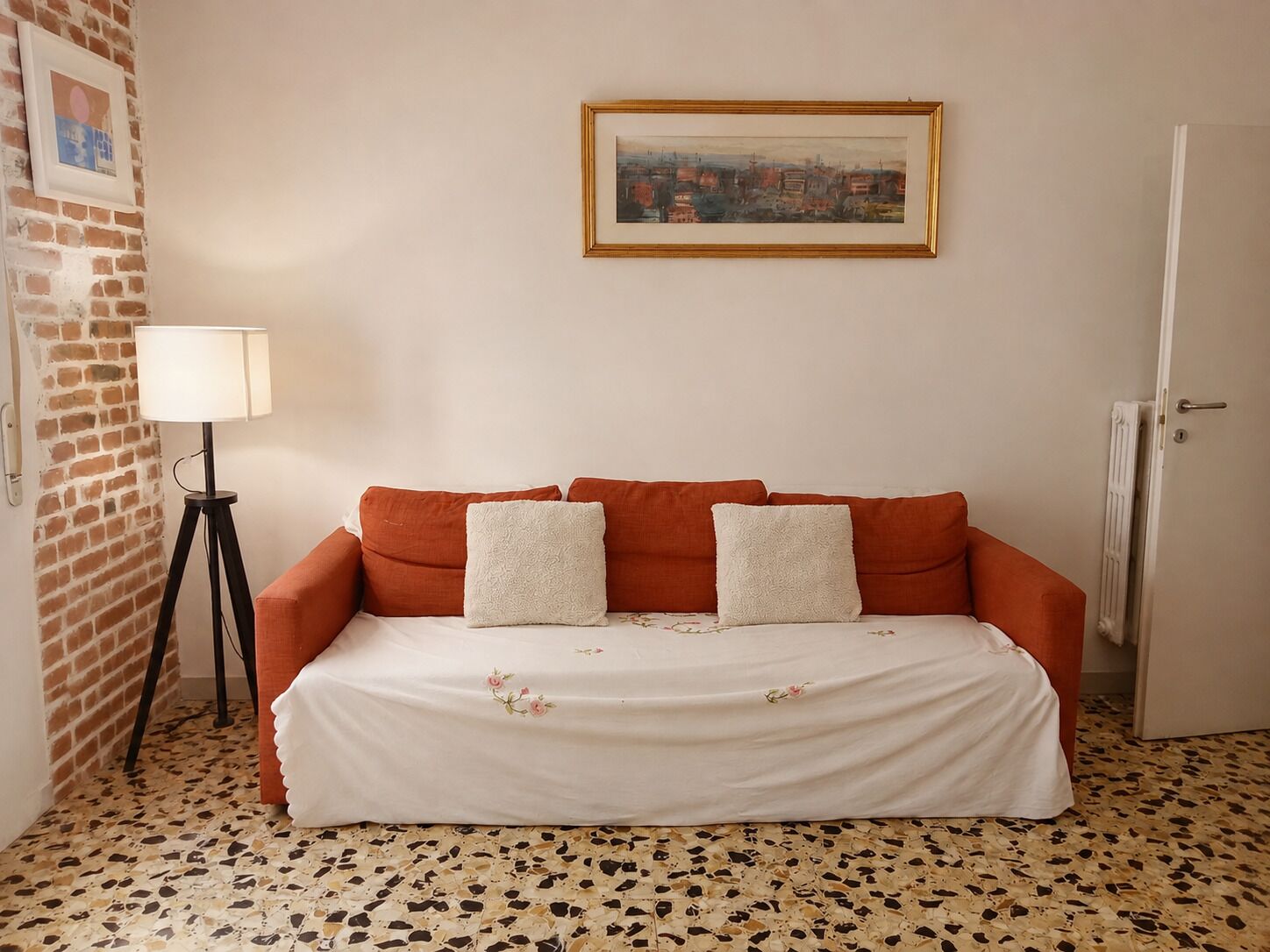 Una stanza con un muro in mattoni, un quadro incorniciato, una lampada da terra e un divano con una coperta a fiori.