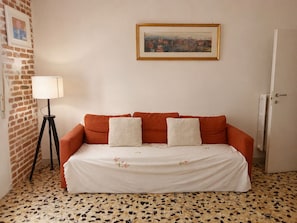 Habitació amb paret de maó, un quadre amb marc, una làmpada de peu i un sofà amb colcha estampada de flors.