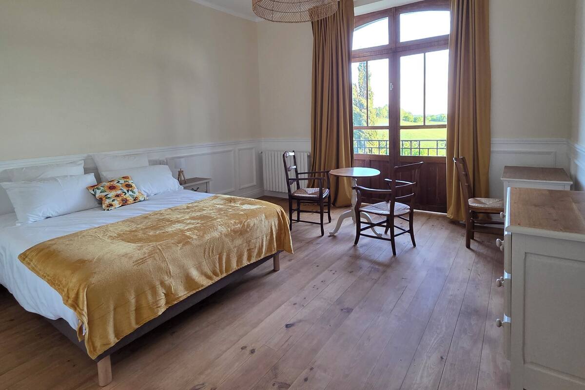 A bedroom with a bed, a table, chairs, a hanging light fixture, and a window with curtains.