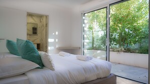 Une chambre moderne avec un grand lit, une porte vitrée donnant sur un balcon et une vue sur la verdure environnante.