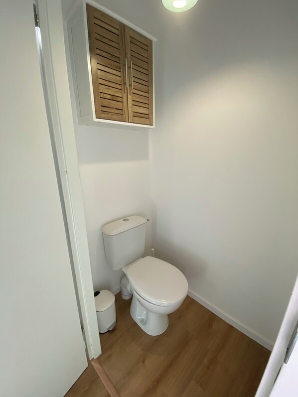 A small, clean bathroom with a toilet, a small trash bin, and a wooden window above the toilet.