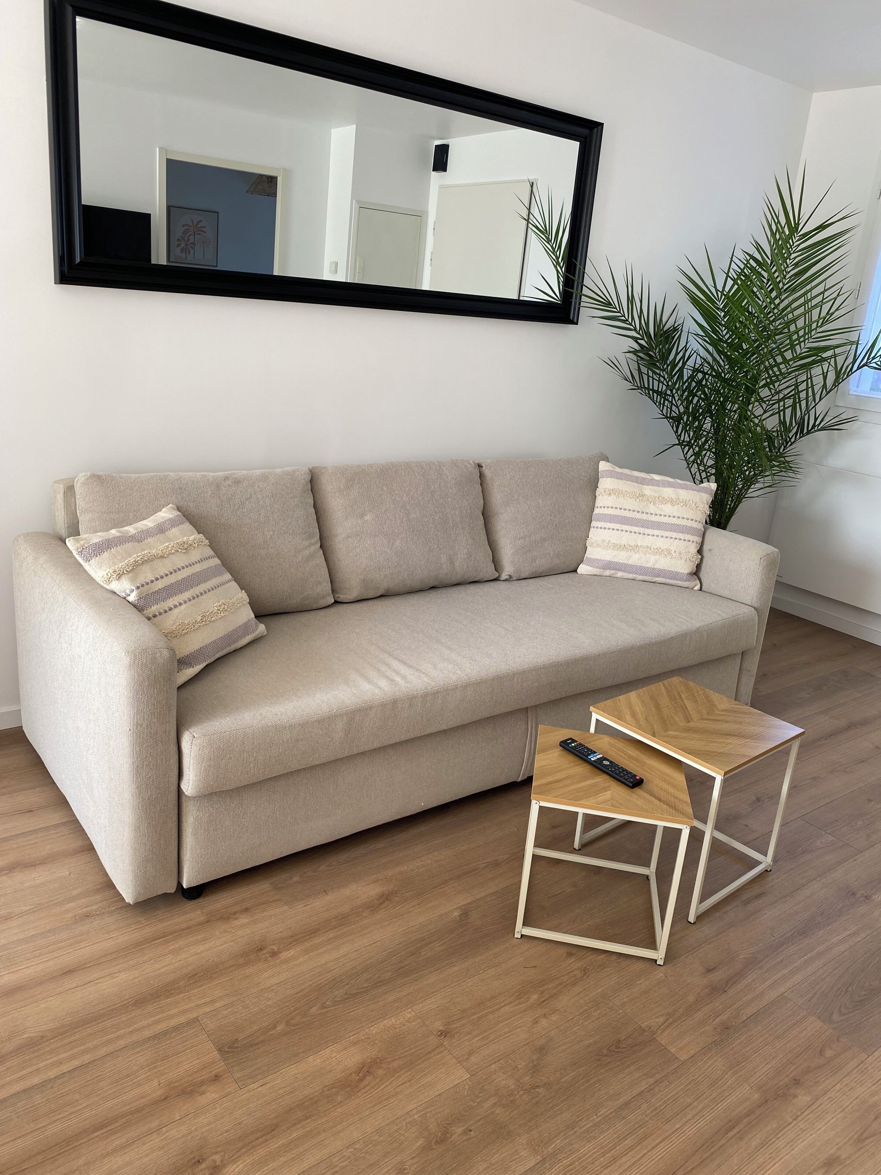 A beige sofa with two pillows, a small wooden side table with a remote control, and a large mirror on the wall.
