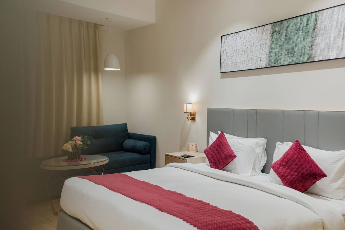 A hotel room with a bed, a nightstand, a lamp, a chair, and a wall-mounted artwork.