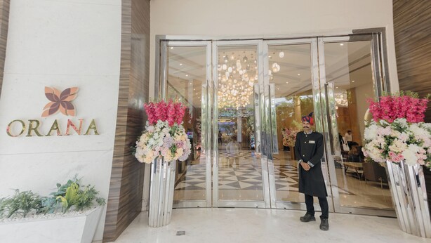 A hotel entrance with a glass door, a receptionist, and floral arrangements. - Orana Hotel & Convention ,Vaishali 1 minute Walk from Vaishali Metro Station. (Ghaziabad)