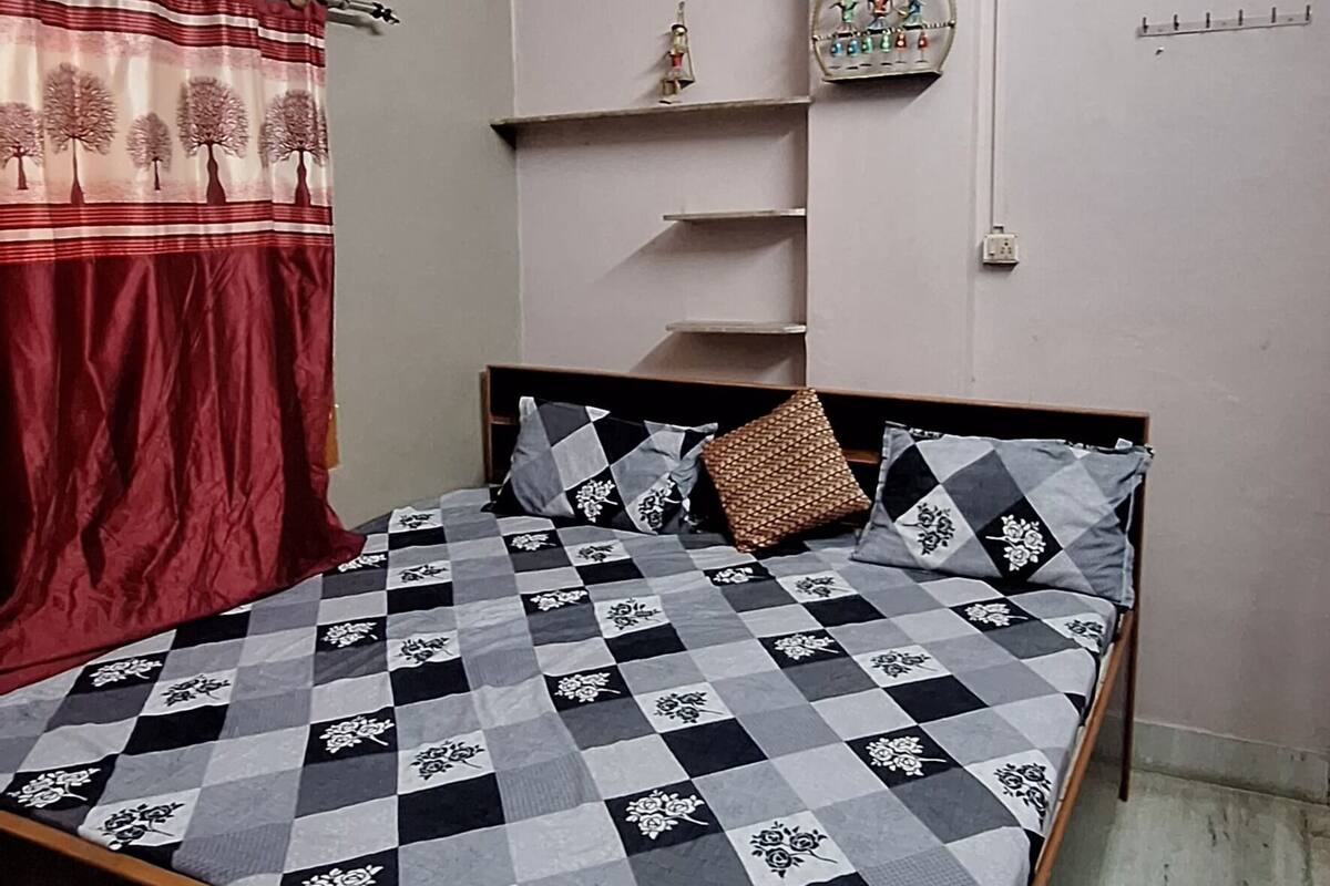 Um quarto com cama, ventoinha no teto, aparelho de ar condicionado, janela com cortinas e prateleira na parede com objetos decorativos.