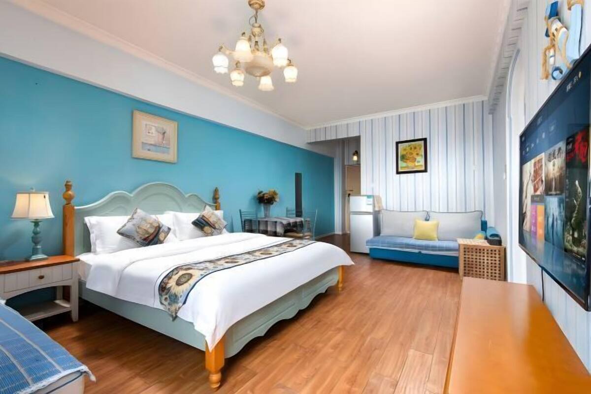 A bedroom with a bed, a desk, a chair, a sofa, a chandelier, and a wall-mounted TV.