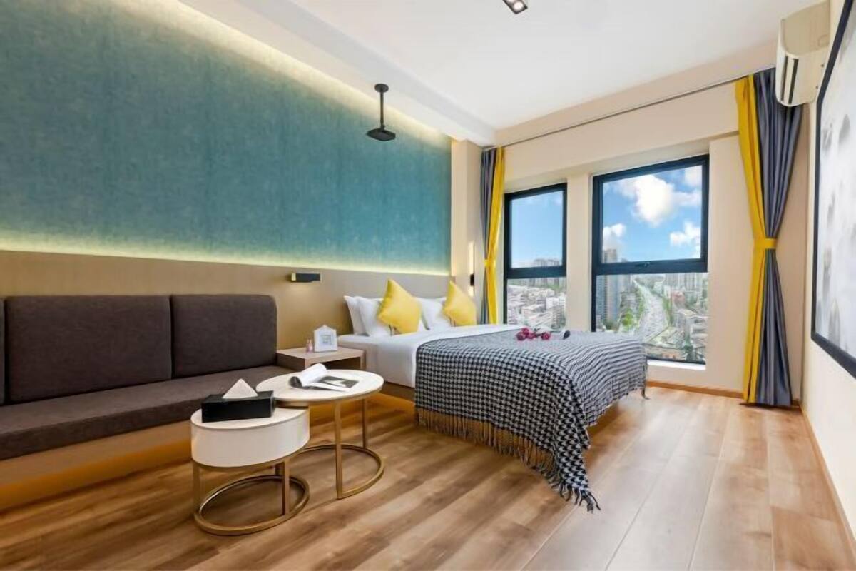A modern hotel room with a sofa, bedside table, bed with a checkered bedspread, and a large window with curtains.